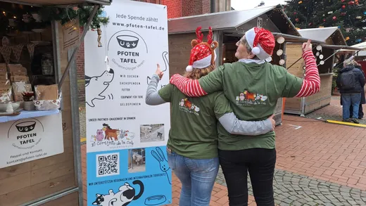 Two female volunteers hugging next to the Pfoten-Tafel association's sign